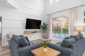 A modern living room with a flat-screen TV, grey sofas, a wooden coffee table, and a large window with a view of a swimming pool.
