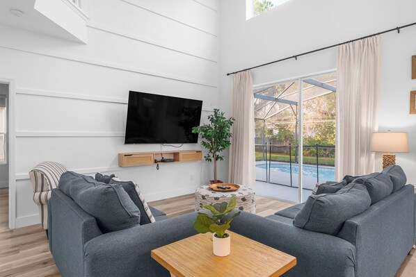 Un salón moderno con televisor de pantalla plana, sofás grises, una mesa de centro de madera y un ventanal con vistas a una piscina.