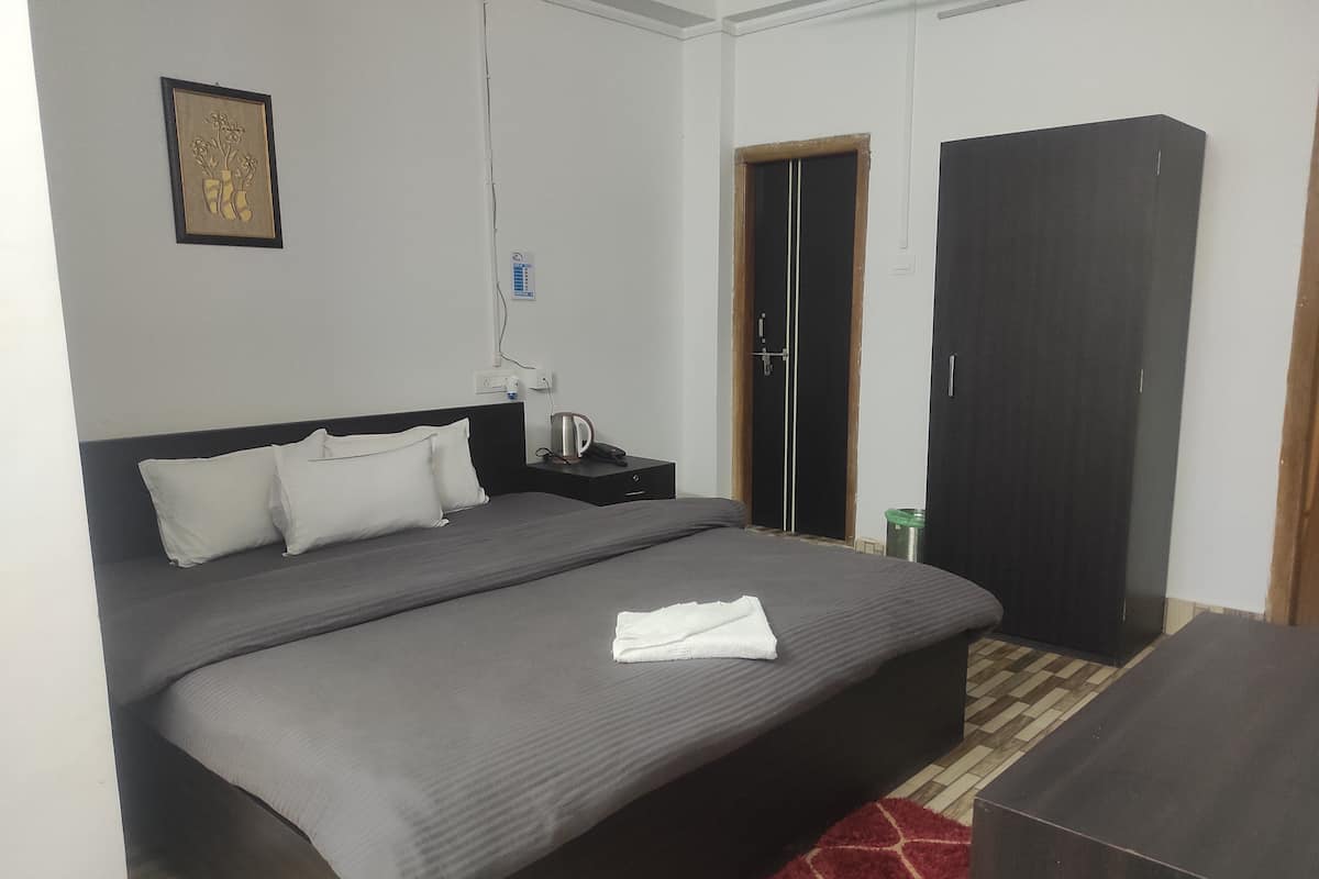 A neatly made bed with a grey bedspread and white pillows, a bedside table with a kettle, a framed picture on the wall, and a wardrobe.