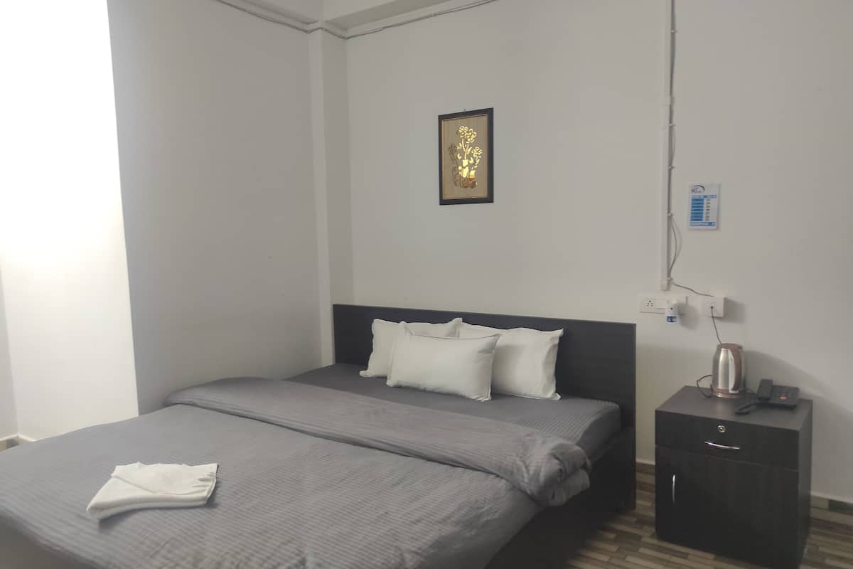 A neatly made bed with grey bedding and white pillows, a small bedside table with a kettle and phone, and a wall-mounted power outlet.