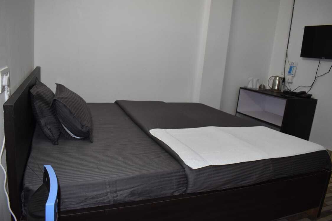 A single bed with a dark mattress and pillows, a bedside table with a lamp, and a wall-mounted TV.