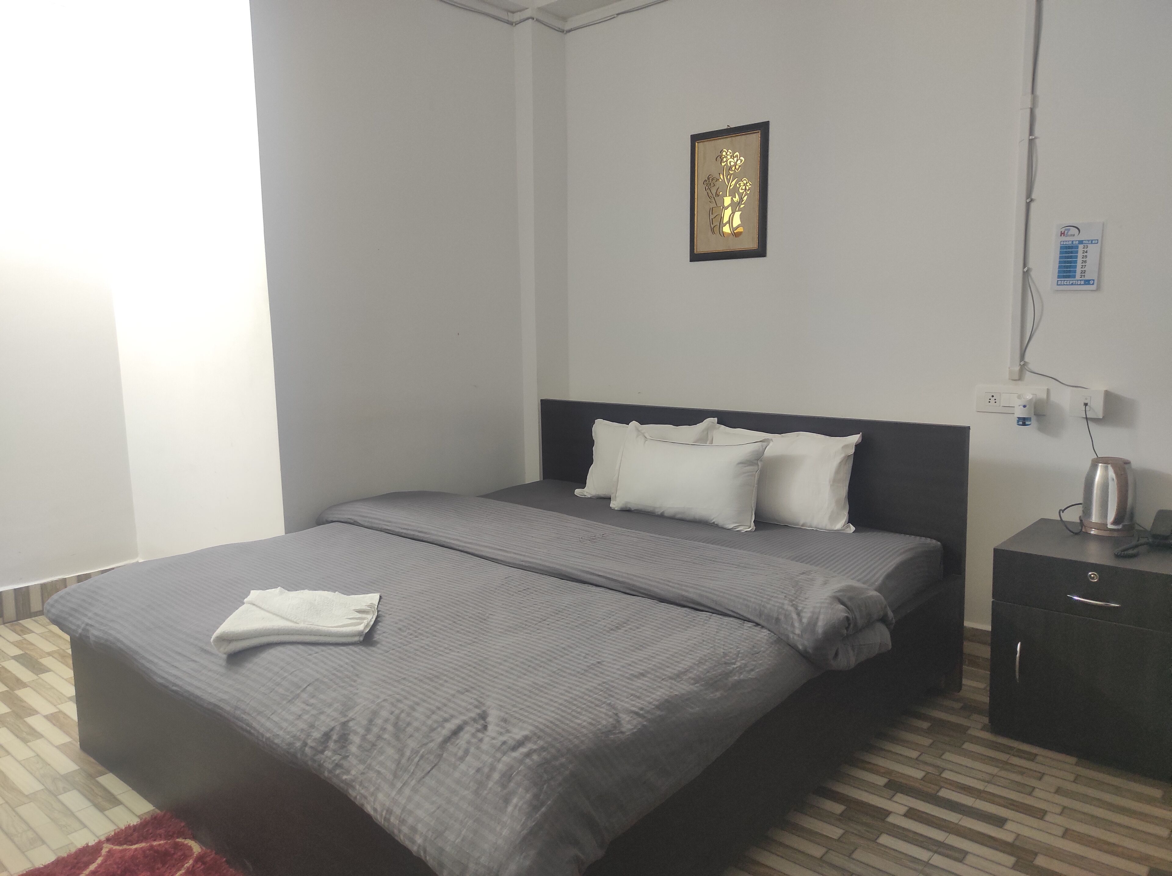 Een net opgemaakt bed met een grijs-wit gestreept dekbed, een donker hoofdbord en witte kussens. Aan de muur hangt een ingelijste foto, een nachtkastje met een waterkoker en een stopcontact.