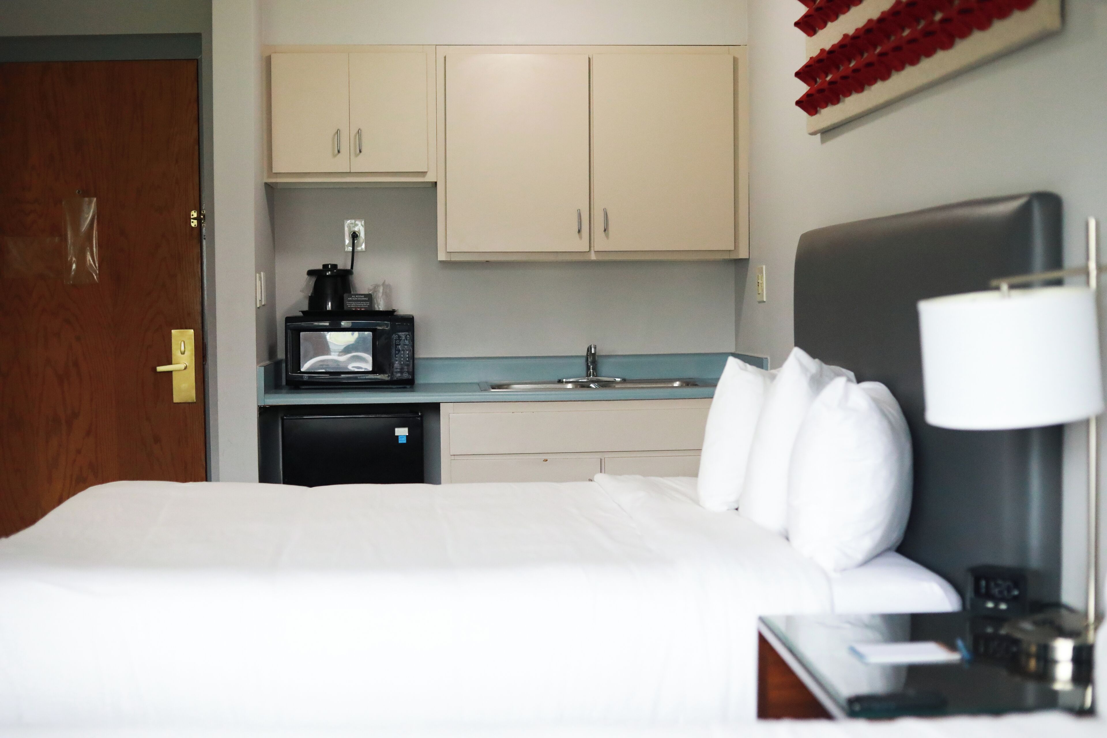 Ein Hotelzimmer mit Bett, Nachttisch mit Lampe, einer kleinen Küchenzeile mit Mikrowelle und Spüle.