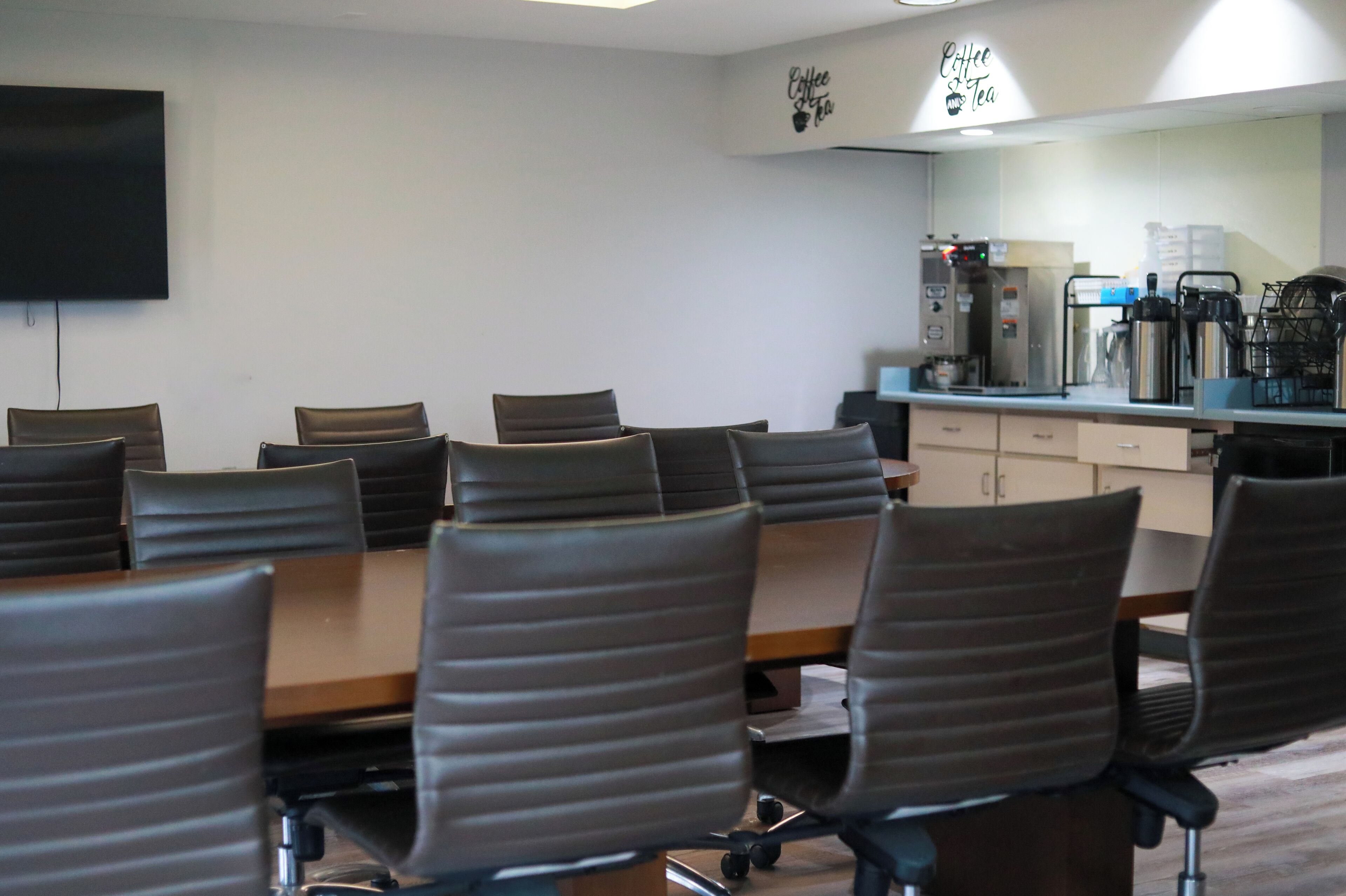 A conference room with a long table and several chairs, a coffee machine, and a TV mounted on the wall.