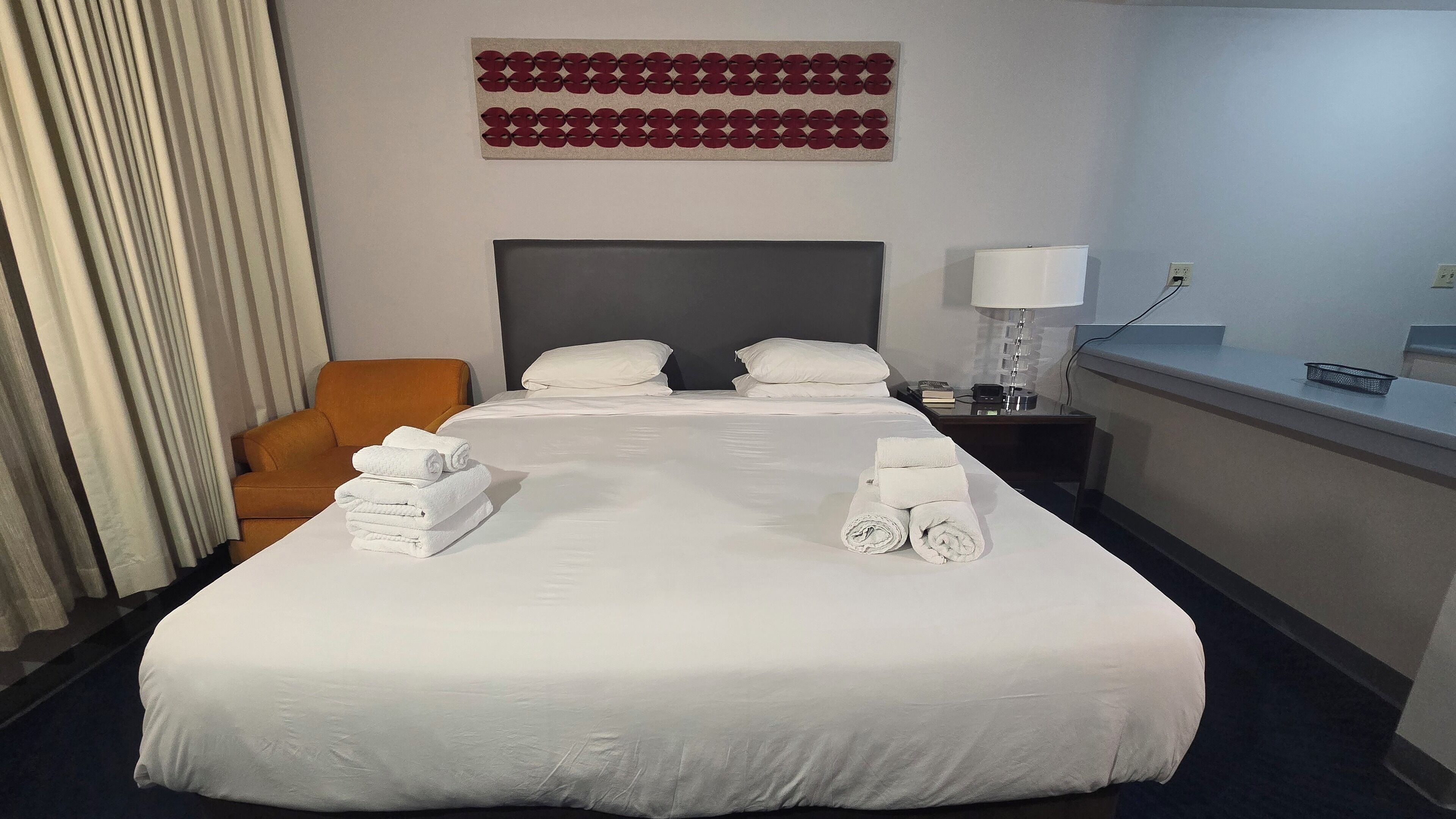 Habitación de hotel con una cama grande, dos almohadas, una mesita de noche con lámpara, una silla y una obra de arte en la pared.