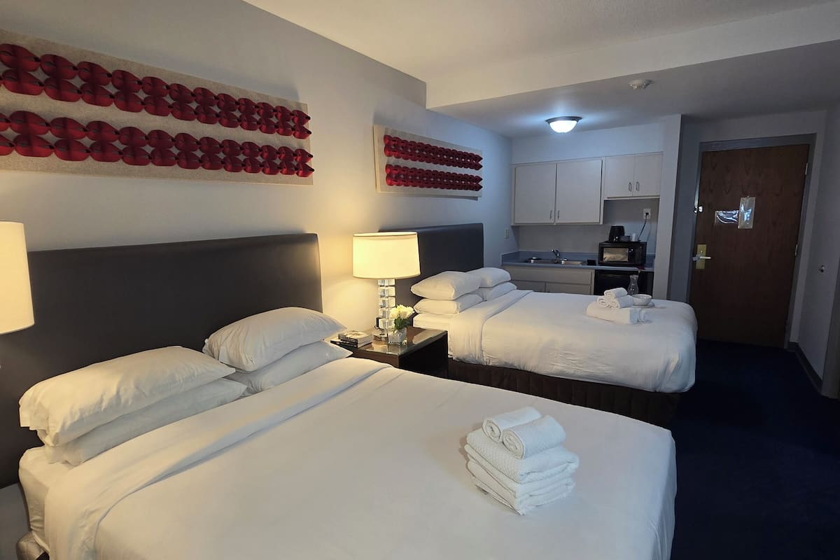 A hotel room with two beds, a desk, a lamp, and a kitchenette.