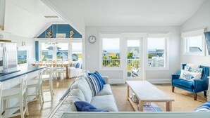 Uma sala ampla com um sofá grande, uma mesa de centro e uma poltrona azul. Há uma cozinha com armários brancos e uma mesa de jantar com cadeiras. O quarto possui janelas grandes com vista para o exterior.