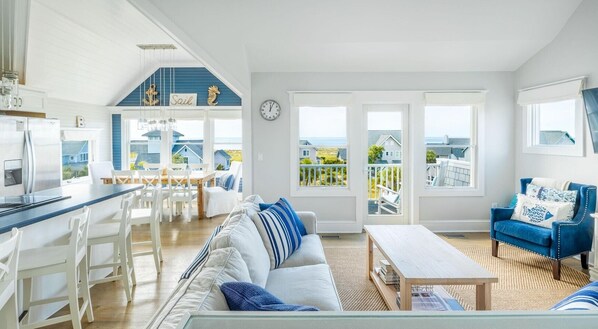 Un vaste espace de vie comprenant un grand canapé, une table basse et un fauteuil bleu. On y trouve également une cuisine avec des meubles blancs et une table à manger avec des chaises. La pièce est dotée de grandes fenêtres offrant une vue sur l’extérieur.