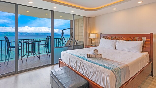 A hotel room with a large bed, a balcony with a view of the sea, a ceiling fan, and a nightstand with a lamp. - Valley Isle Resort #1107 - Vacation West Maui - Beachfront Oasis (Lahaina)