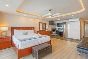 A modern bedroom with a wooden bed, a nightstand, a bench, a ceiling fan, and a kitchenette in the background.