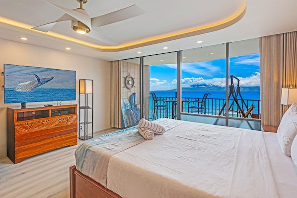A bedroom with a large bed, a TV, a wooden dresser, and a balcony with a view of the sea.