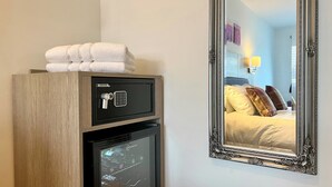 A minibar with a safe and a refrigerator, a mirror reflecting a bed with pillows, and a wall-mounted lamp. - Fyne Rooms - Arran Superior King - Bed & Breakfast (Tighnabruaich)