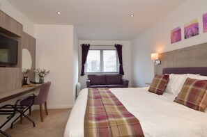 Ein Hotelzimmer mit Bett, Schreibtisch mit Stuhl, Fernseher, Fenster mit Vorhängen und einer Wandlampe.