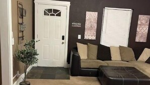 A living room with a beige carpet, a black leather sofa, a black ottoman, a white door, and a small potted plant.