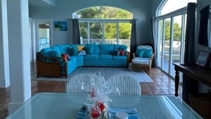 A spacious living room with a blue sofa, a glass coffee table, and a view of the outdoors through large windows. - Villa Mangorosa (St Thomas)