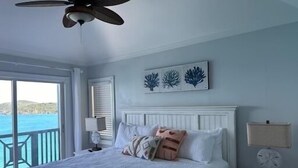A bedroom with a bed, a ceiling fan, a nightstand, a lamp, and a view of the sea. - Villa Mangorosa (St Thomas)