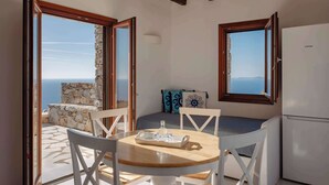 Una habitación con pared de piedra, techo de madera, una mesa redonda de madera con sillas, un refrigerador blanco y vista al mar a través de ventanas abiertas.