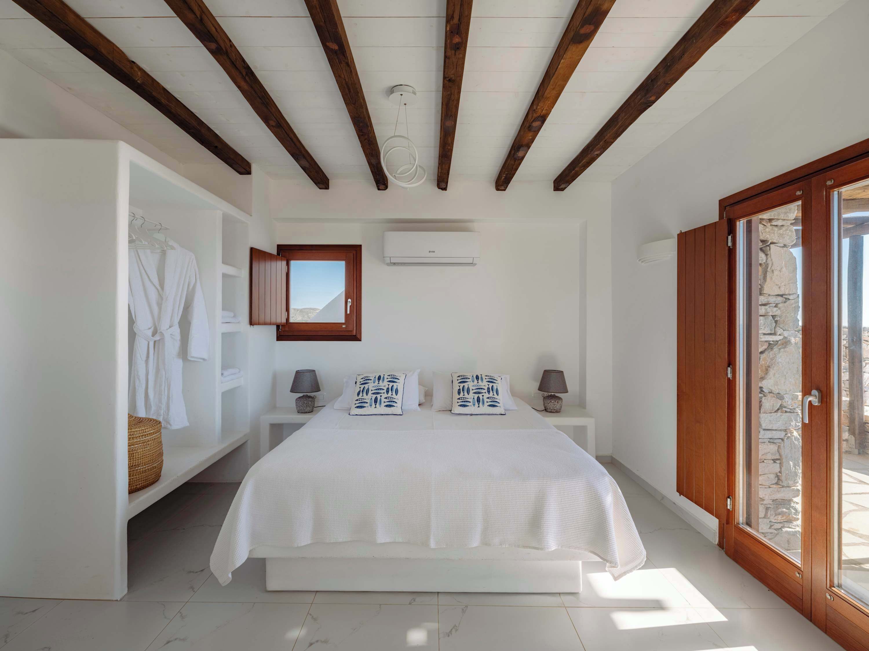 A bedroom with a large bed, wooden beams, a stone wall, and a view through a glass door.