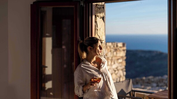 Una persona de pie junto a un ventanal con vista al mar y una pared de piedra.