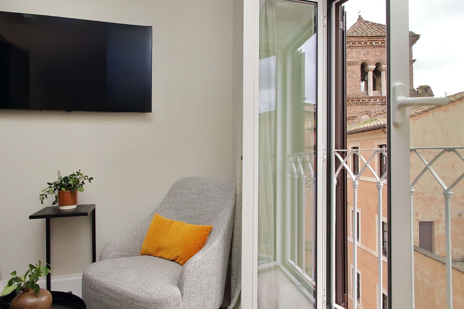 Ein Balkon mit einem grauen Sessel, einem gelben Kissen, einer Topfpflanze und einem an der Wand befestigten Fernseher.