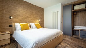 A modern bedroom with a wooden bed, a nightstand, a built-in wardrobe, and a wall-mounted light fixture. - Three-room apartment with garden (Pont-Saint-Martin)