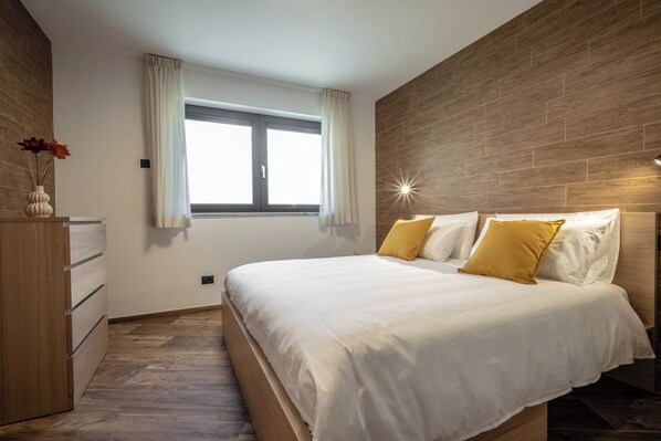 A modern bedroom with a large bed, a wooden dresser, and a window with curtains.