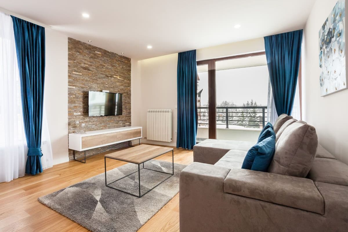 Ein modernes Wohnzimmer mit einer Steinwand, einem Flachbildfernseher, einer Couch mit Kissen und Blick ins Freie.