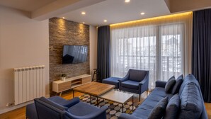 A modern living room with a stone accent wall, a flat-screen TV, a sofa, a coffee table, and a large window with sheer curtains. - Apartments by Bor (Zlatibor)
