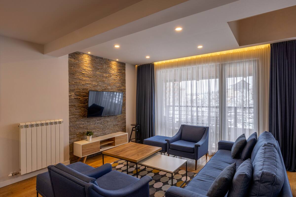 Ein modernes Wohnzimmer mit einer Wand aus Stein, einem Flachbildfernseher, einer Couch, einem Couchtisch und einem großen Fenster mit durchsichtigen Vorhängen.