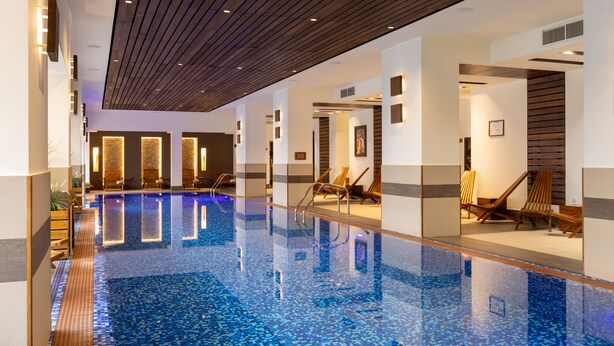 A large indoor swimming pool with clear blue water, wooden ceiling, and lounge chairs along the sides. - Apartments by Bor (Zlatibor)