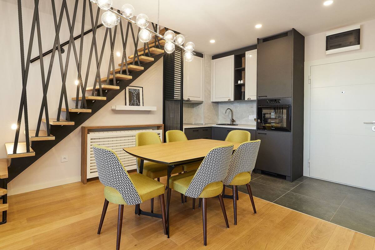 Eine moderne Küche mit integriertem Backofen, ein Essbereich mit Holztisch und gemusterten Stühlen sowie eine Treppe mit einer außergewöhnlichen Leuchte.