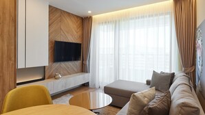 A modern living room with a flat-screen TV mounted on a wooden wall, a sofa with patterned cushions, a coffee table, and sheer curtains on the windows. - Apartments by Bor (Zlatibor)