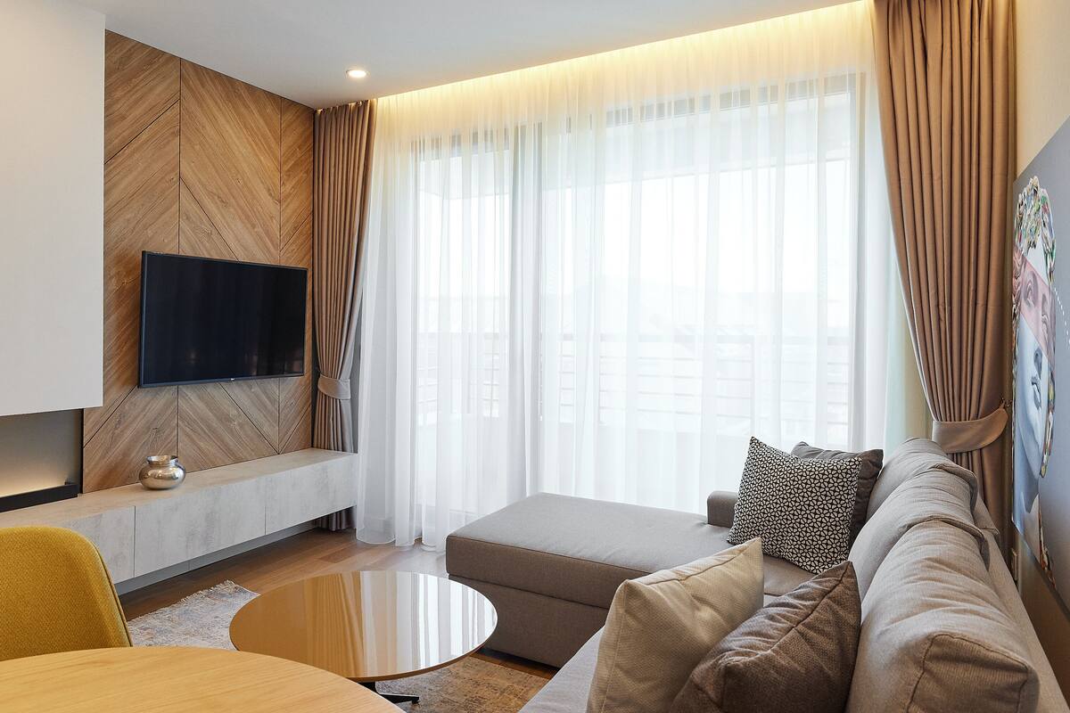 Ein modernes Wohnzimmer mit einem an einer Holzwand montierten Flachbildfernseher, einer Couch mit gemusterten Kissen, einem Couchtisch und durchsichtigen Vorhängen an den Fenstern.