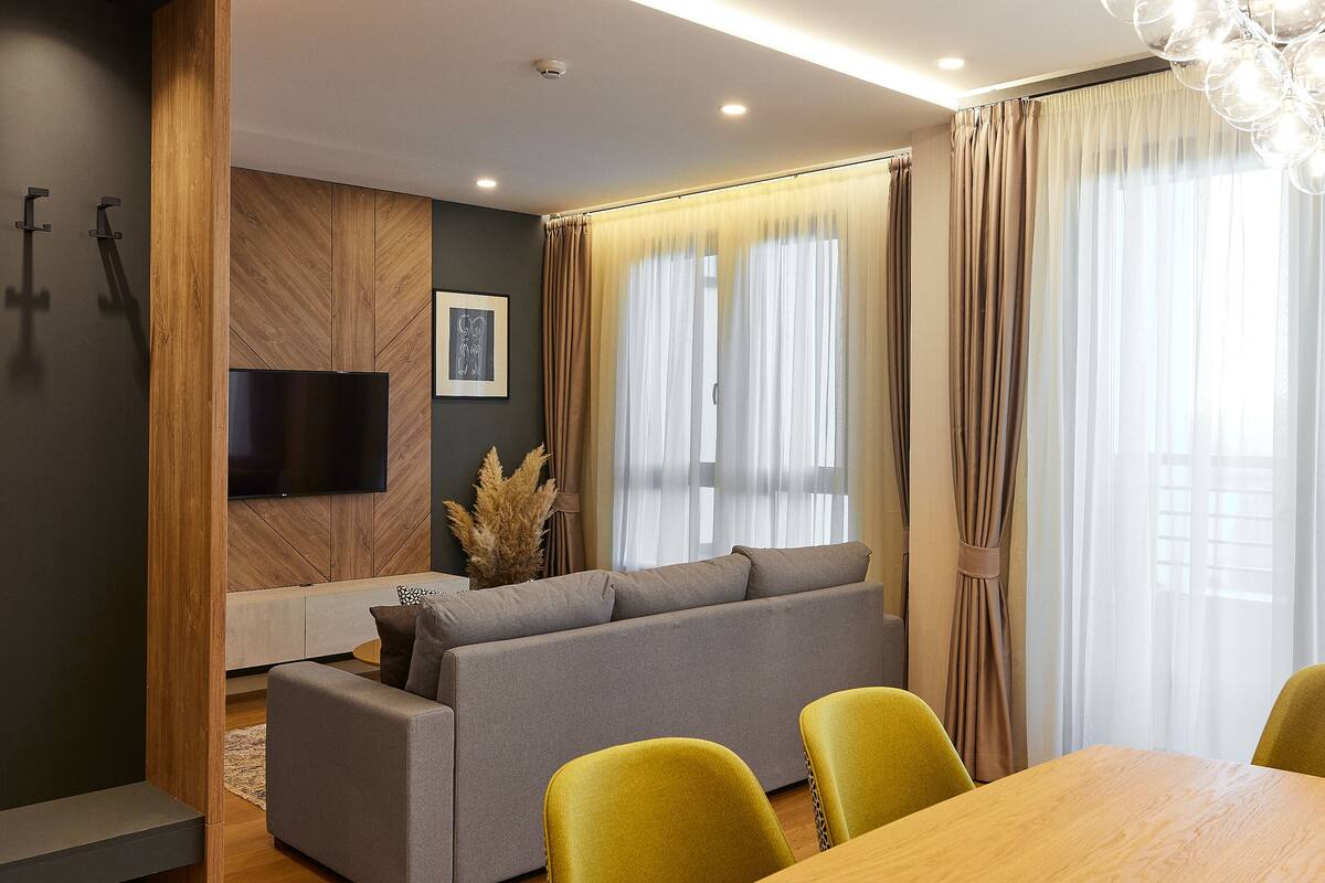 Ein modernes Wohnzimmer mit grauem Sofa, gelben Sesseln, einem Holzesstisch und einem Flachbildfernseher, der an einer Wand aus Holzpaneelen befestigt ist.