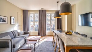 A modern living room with a sofa, coffee table, dining table, and chairs. There is a large window with curtains, a flat-screen TV, and a balcony view.
