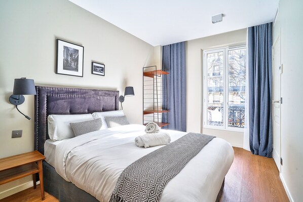 A neatly made bed with a patterned throw, a bedside table with a lamp, and a window with curtains.