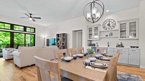 Un soggiorno moderno con zona pranzo, una ventola a soffitto, un lampadario e una cucina con pensili bianchi e un orologio.