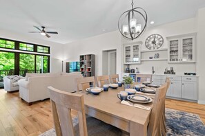 Ein modernes Wohnzimmer mit Essbereich, Deckenventilator, einer Kandelaberleuchte und einer Küche mit weißen Schränken und einer Uhr.