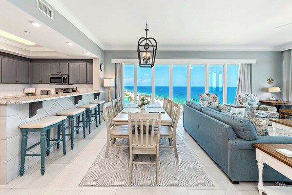 Amplia sala de estar con un gran ventanal con vistas al océano, una cocina con taburetes de bar y una mesa de comedor con sillas.