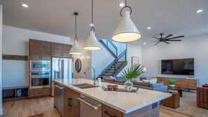 Een moderne keuken met een centraal eiland, roestvrijstalen apparatuur, hanglampen en een trap die leidt naar een woonruimte met een televisie en zithoek.