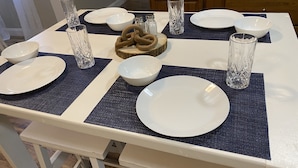 Een eettafel gedekt met witte borden, kommen en helder glaswerk, met een blauwe placemat en een houten bestektafelstuk.