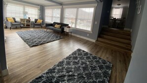 Een ruime woonkamer met houten vloeren, een grijze bank, twee fauteuils en een groot vloerkleed.