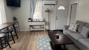 Ein modernes Wohnzimmer mit Holzboden, einem grauen Sofa, einem Couchtisch, einem Schreibtisch mit Stuhl, einem an der Wand befestigten Fernseher und einem Fenster mit Vorhängen.