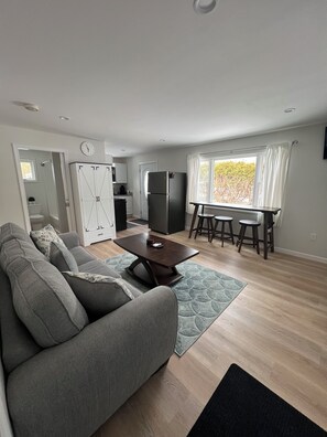 Un salon moderne avec un canapé gris, une table basse en bois et un coin repas comprenant une table et des tabourets.
