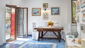 A dining area with a wooden table, white chairs, a vase of flowers, and a tiled floor. - Olivegrove Holiday House (Massalubrense)