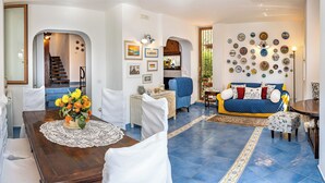 A spacious living room with a blue tiled floor, a wooden dining table with a lace tablecloth, and a blue sofa with patterned cushions. - Olivegrove Holiday House (Massalubrense)