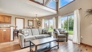 A spacious living area with a sofa set, a coffee table, and a dining table. There is a kitchen area with wooden cabinets and a window overlooking a garden.