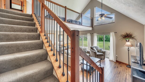 A spacious living area with a wooden staircase, a ceiling fan, and a large window offering a view of the outdoors.