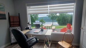 A room with a desk, a chair, a laptop, a grill, and a view of a lake and trees. - Lochaber Lake View Cottage (Antigonish County)
