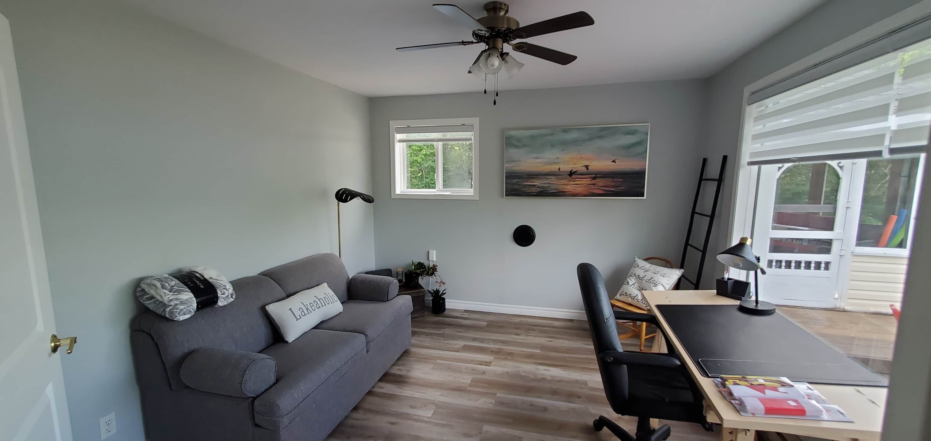 Et værelse med en grå sofa, en sort kontorstol, et skrivebord med lampe, en loftventilator og et vindue med persienner.